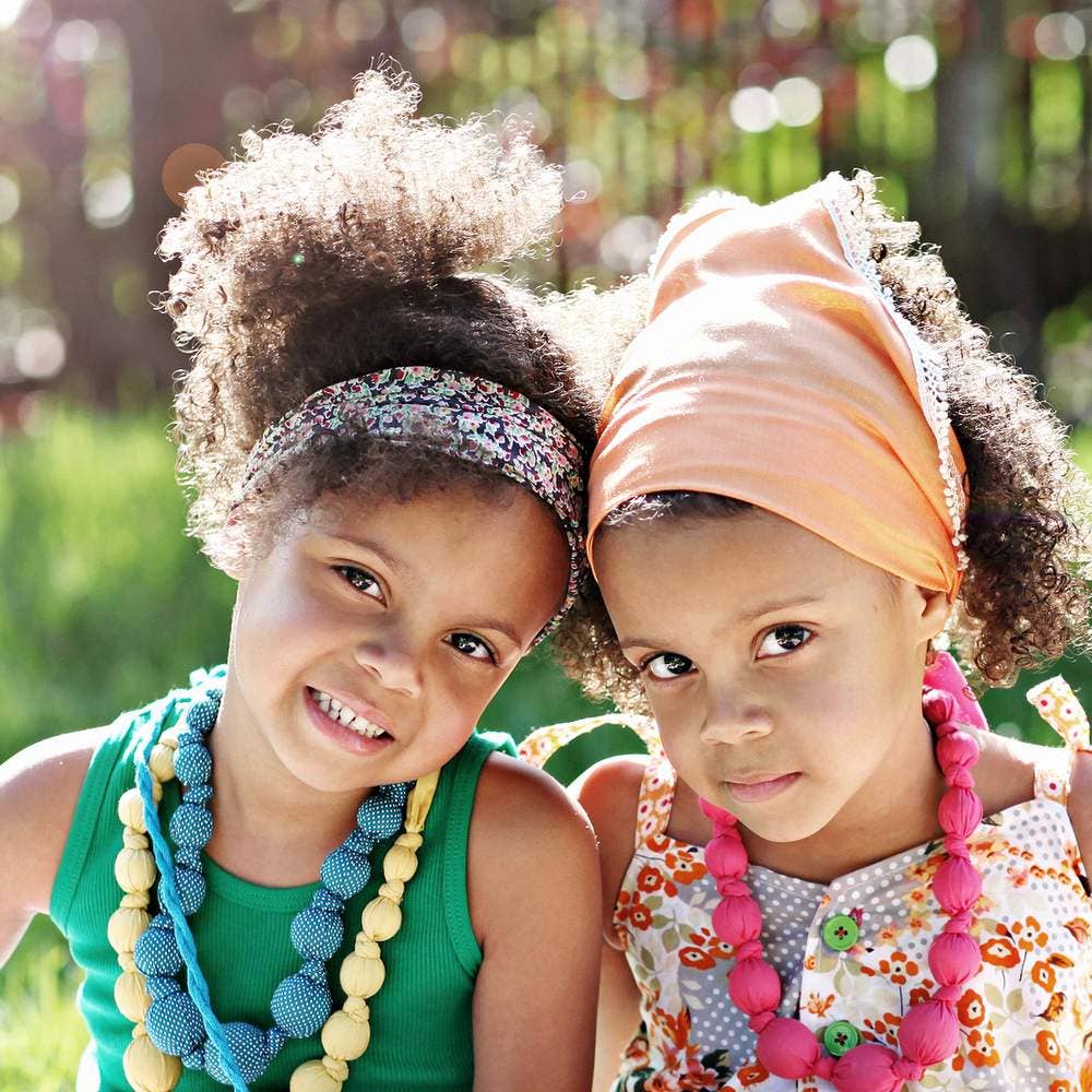 Two young girls wearing colorful headbands and necklaces, sitting outdoors.